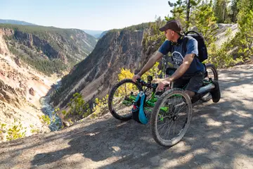 A person in an off-road wheelchair on a forest trail, with a companion walking beside them