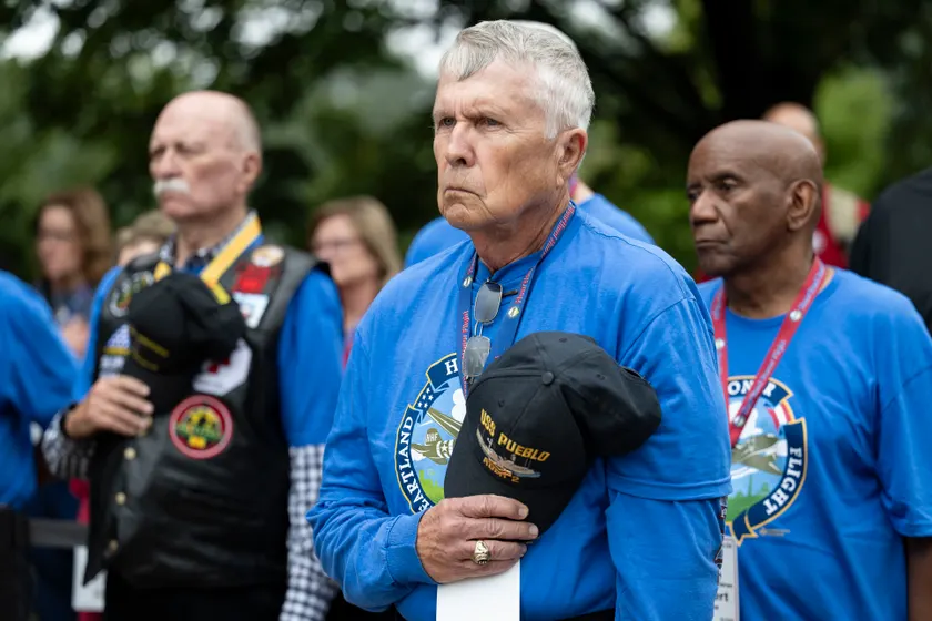 Three older men in blue honor-flight shirts stand together outdoors, one holding a black cap