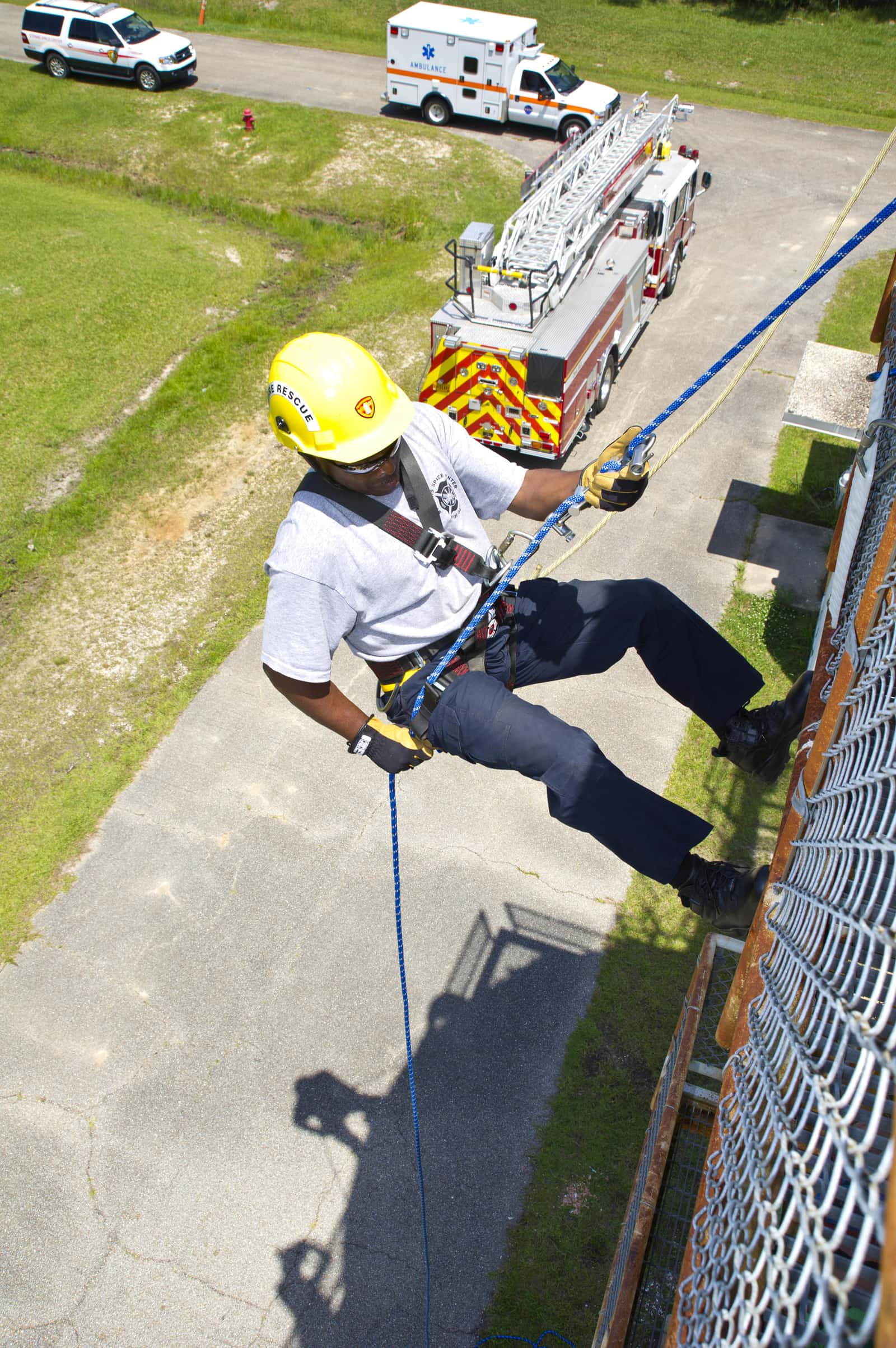 A firefighter in a yellow helmet rappels down a tower on a blue rope, with a ladder truck parked below