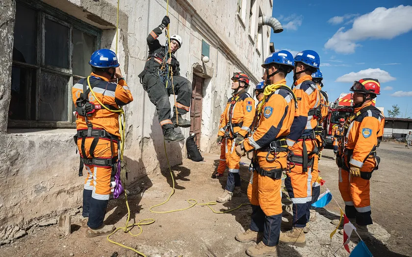 Rescuers in helmets and harnesses watch a teammate climb a rope beside a building