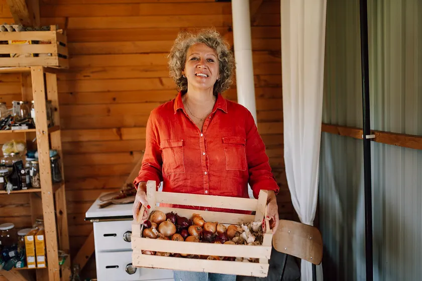 A smiling woman in a red shirt holding a wooden crate of onions, garlic, and other vegetables