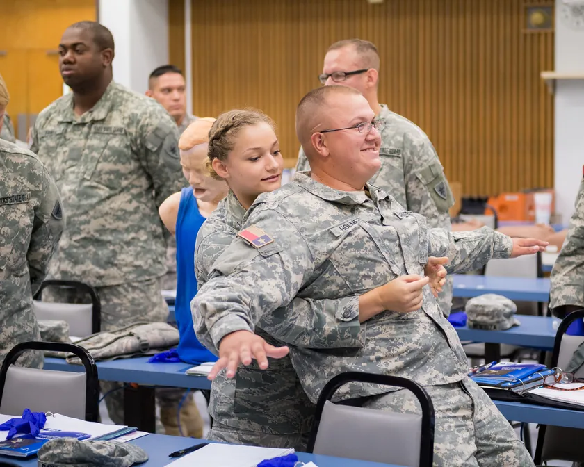 Two people in camouflage uniforms practice abdominal thrusts in a classroom while other trainees watch