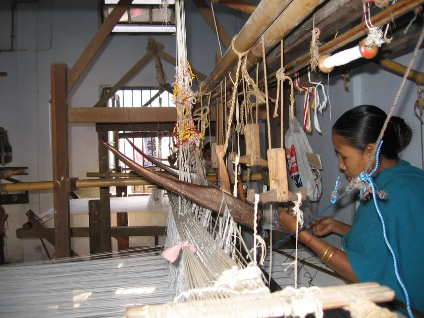 A woman in a teal shirt working at a wooden handloom strung with white threads