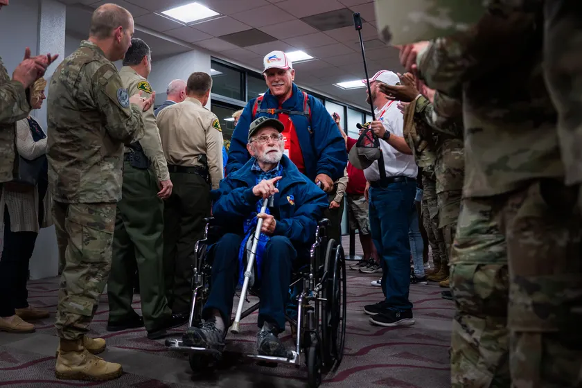 A smiling veteran in a wheelchair moves through an airport hallway while people in uniform applaud