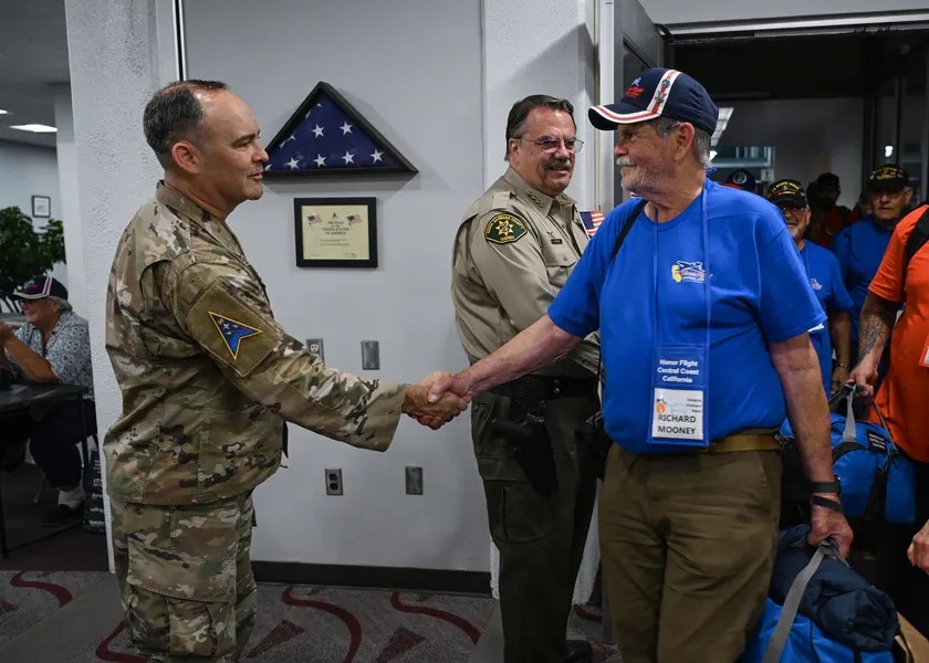 A service member in camouflage shaking hands with an older veteran in a blue Honor Flight shirt