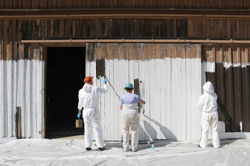 Three people in work clothes painting the wooden doors of a weathered barn