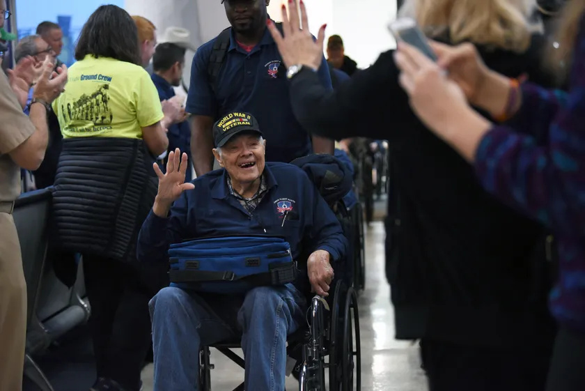 An older man in a wheelchair and a World War II veteran cap smiles and waves while people clap in an airport hallway