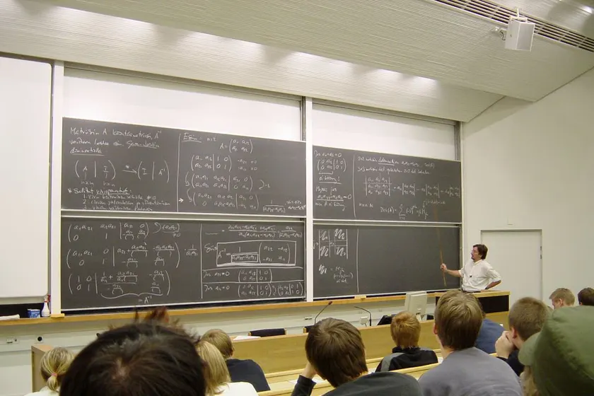 University students seated in a lecture hall, seen mostly from behind, facing a whiteboard