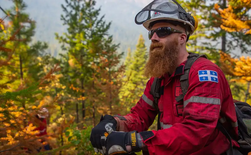 A bearded firefighter in a red jacket and helmet standing in a forest
