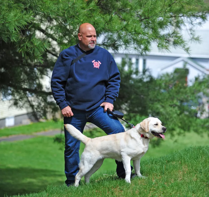A man in a navy fire-department sweatshirt holds a leash while a yellow Labrador stands on the grass beside him