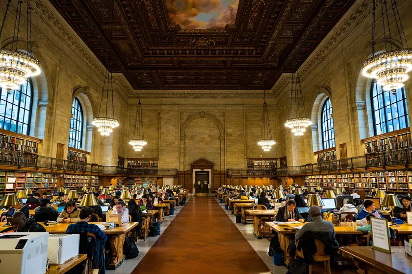 People studying at wooden tables under chandeliers in a large library reading room
