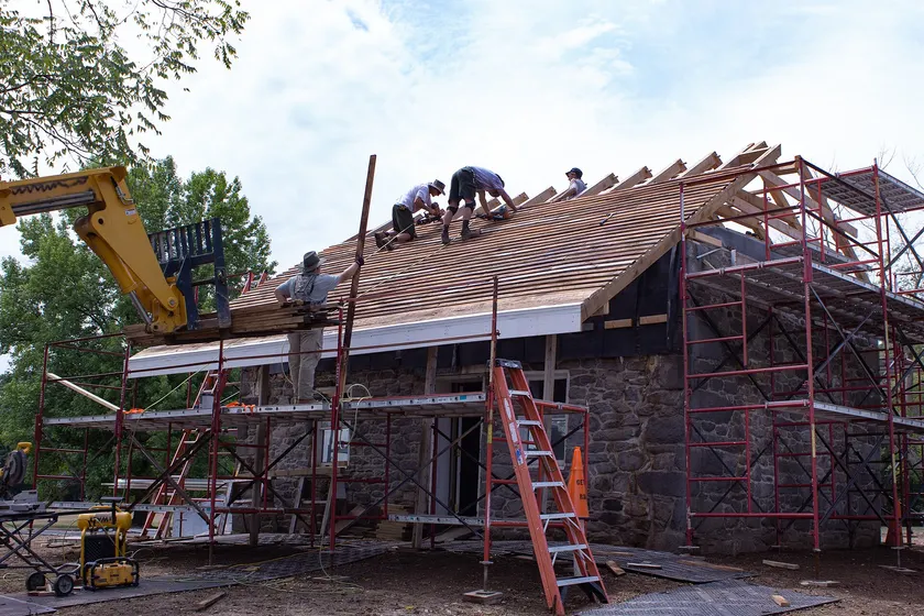 Workers on a steep roof while a machine lifts a bundle of cedar shingles