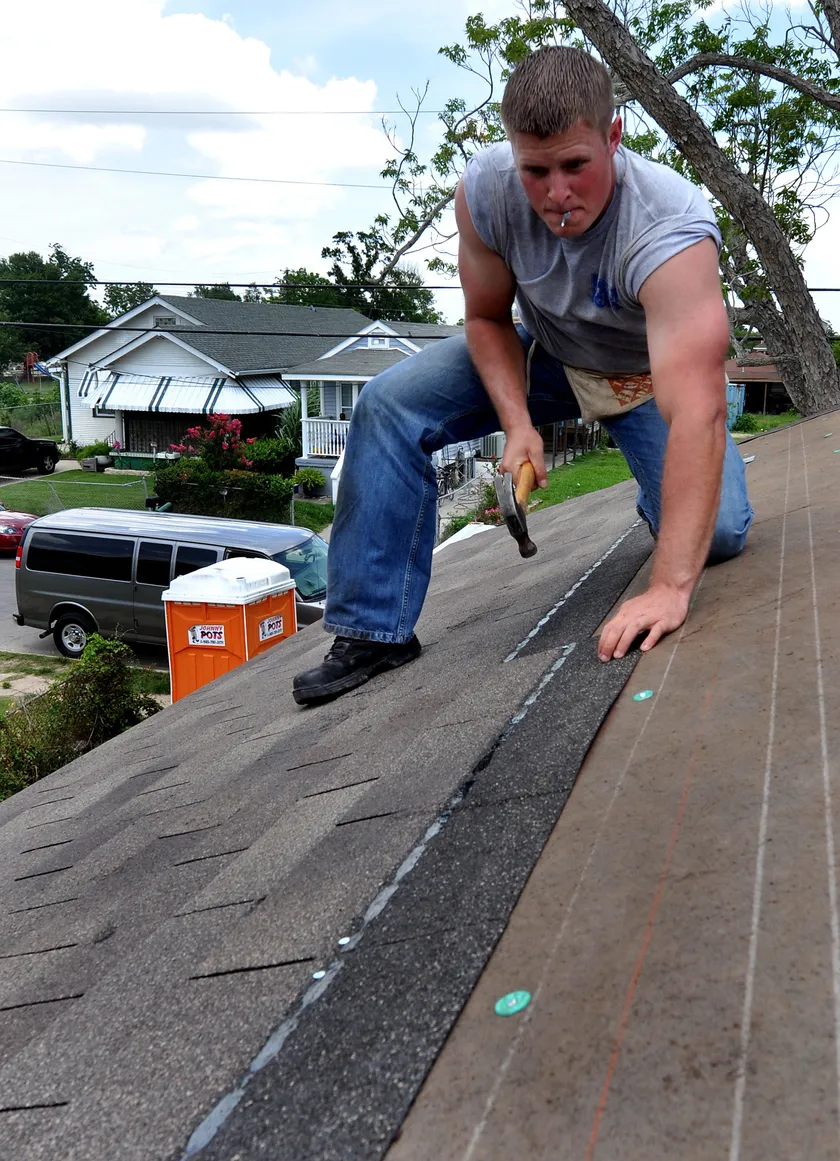A man kneels on a roof and holds a hammer over the decking