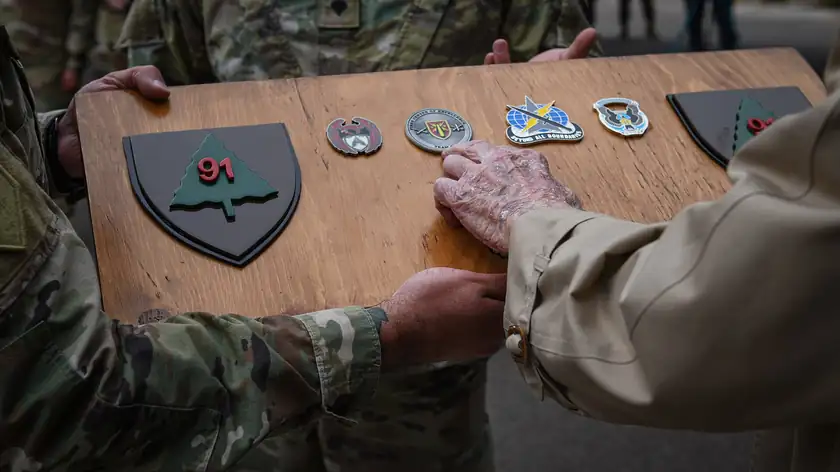 An older person's hand touching coins on a wooden plaque held by people in uniform