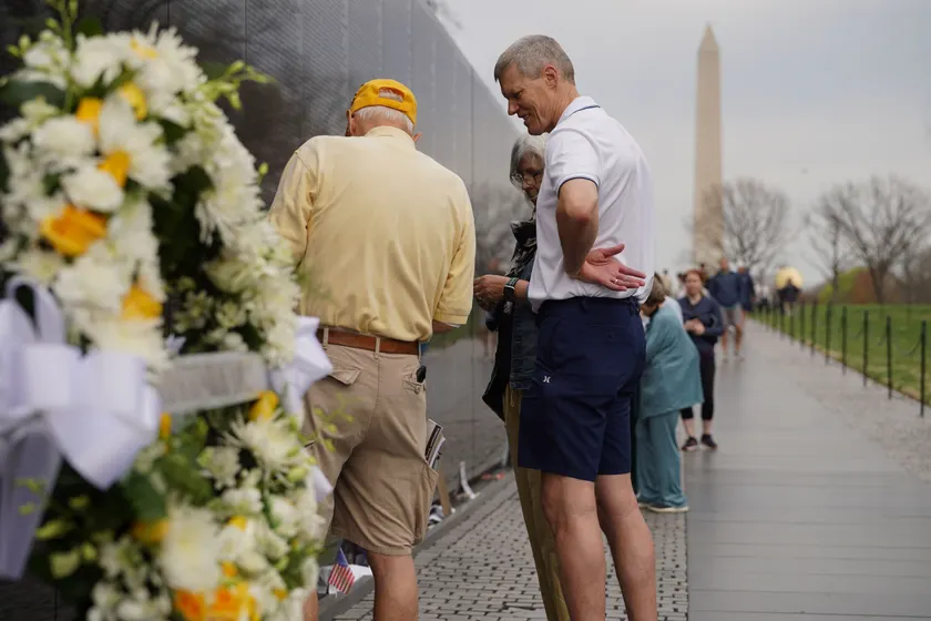 People stand beside a flower wreath at the Vietnam Veterans Memorial, with the Washington Monument in the distance