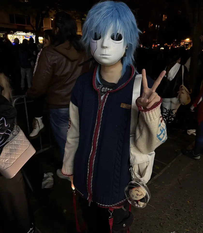 A person in a blue wig and a white mask making a peace sign at a nighttime costume parade