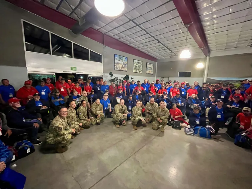 Service members in camouflage kneel in front of a large group of veterans wearing blue and red shirts inside an airport hall