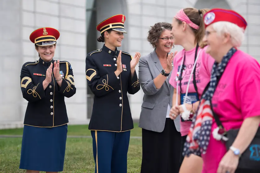 Soldiers in dress uniforms applauding women in honor-flight shirts outside a stone memorial