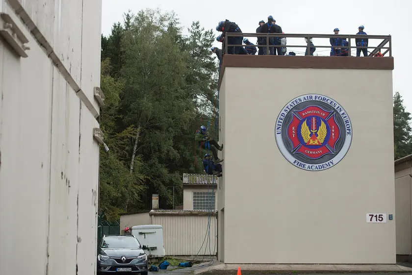 Firefighters in helmets lowering a person on ropes beside a training tower