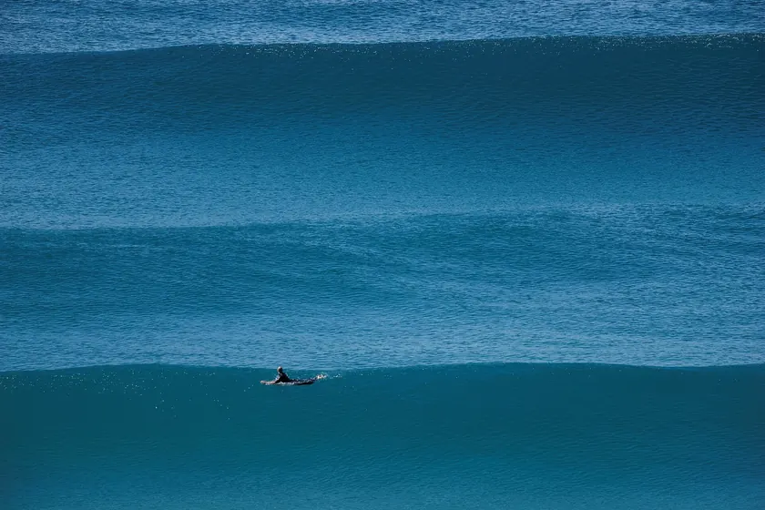 A surfer in a full black wetsuit paddling a white surfboard through choppy waves