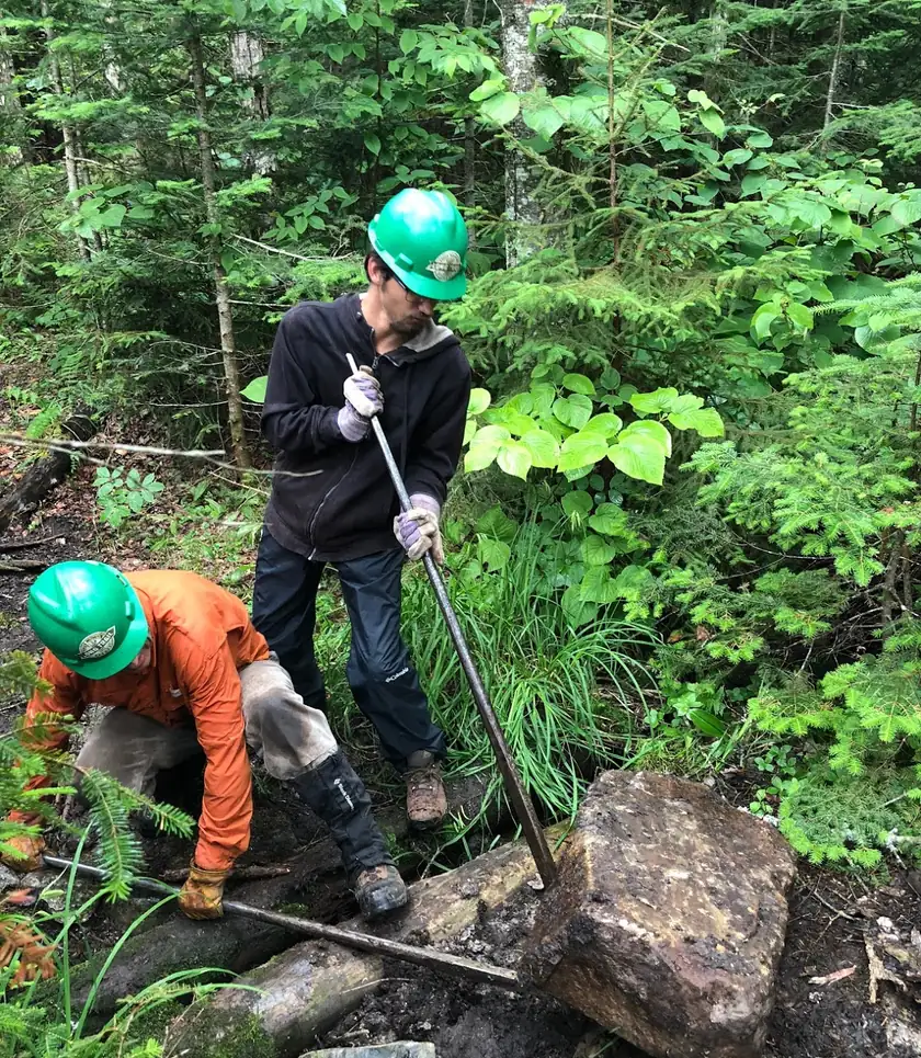 Two trail workers in green helmets moving a rock on a forest path