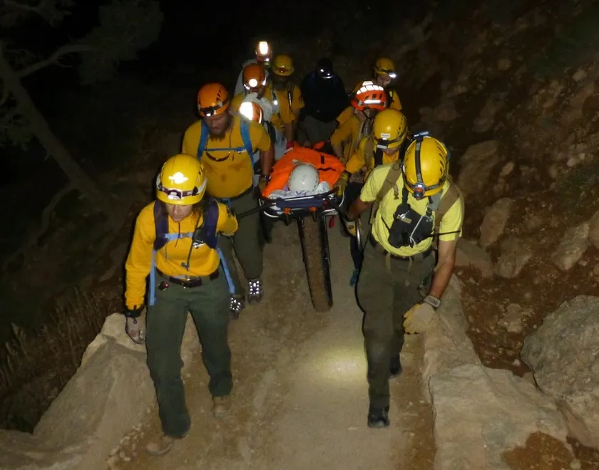 A team in yellow helmets carries a person on a rescue litter up a rocky trail at night
