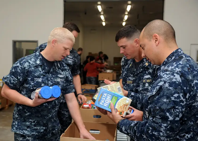 Three sailors in uniform sorting and packing food at a food bank table