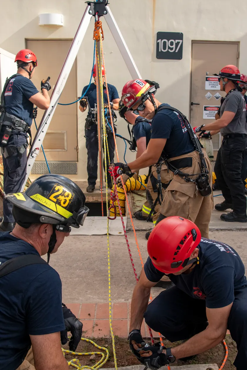 Firefighters in helmets handle ropes on a metal tripod set over an opening in a concrete pad