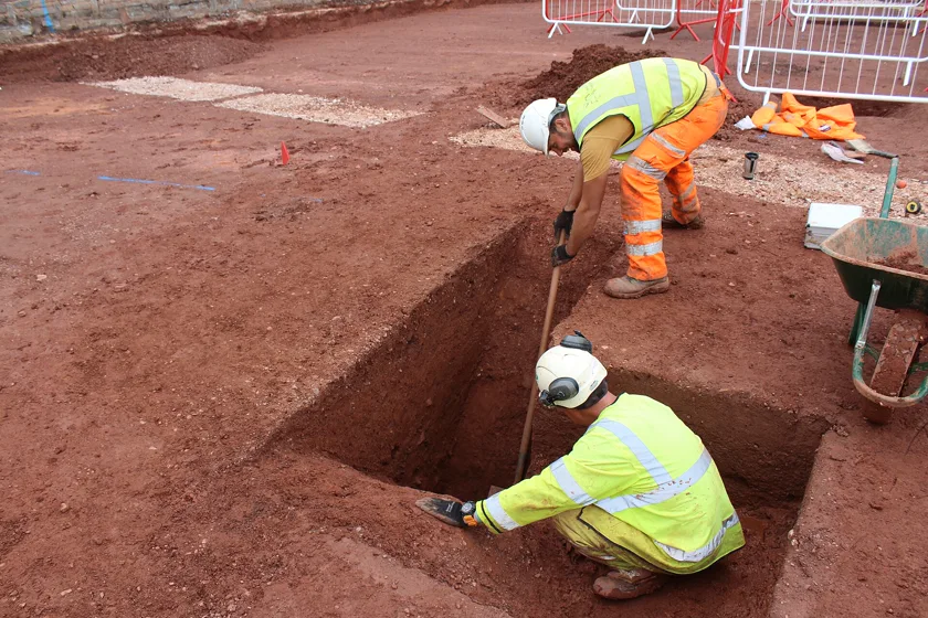 Two archaeologists in high-visibility clothing excavating a deep trench
