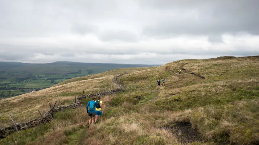 Two runners with backpacks following a grassy trail beside a stone wall on an open hill
