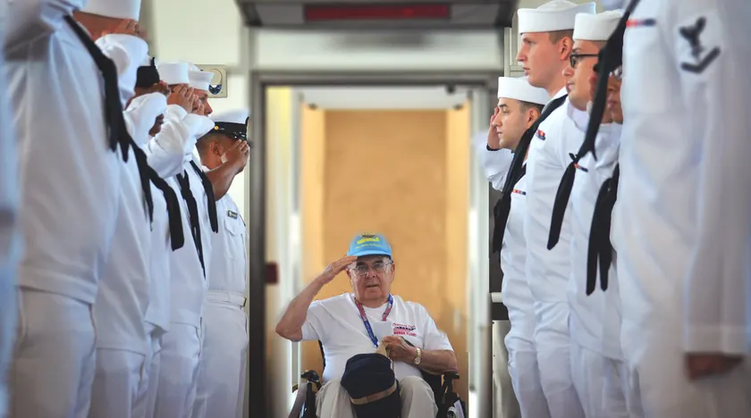 Sailors in white uniforms saluting a veteran in a wheelchair who is saluting back