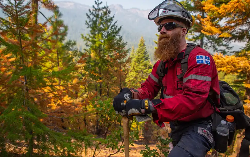 A firefighter in a red jacket and helmet resting a hand on a tool in the woods