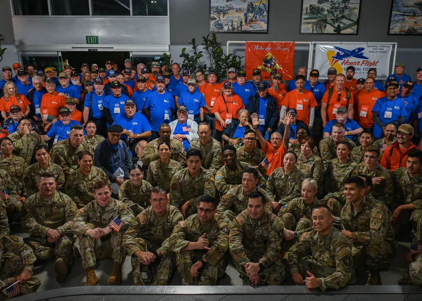A large group of veterans in bright shirts and service members in camouflage pose together indoors, some holding small American flags