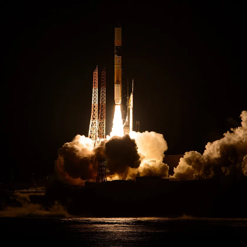 A rocket climbing into a bright sky with a long exhaust plume above a shoreline