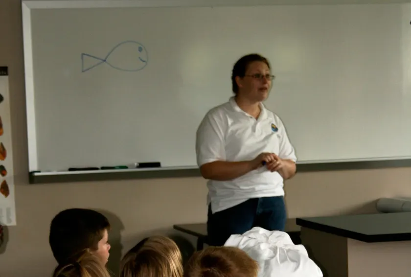 A teacher speaking to students seated at desks, seen mostly from behind