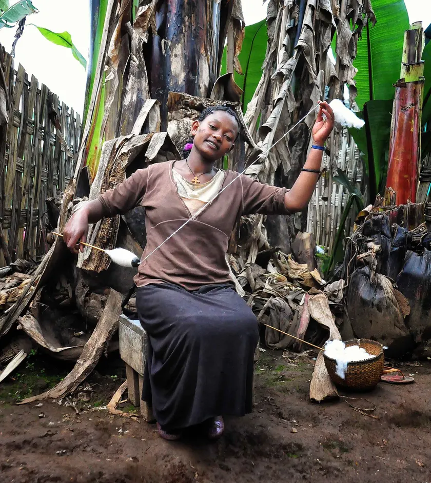 A woman seated outdoors, spinning white fiber between two hand spindles