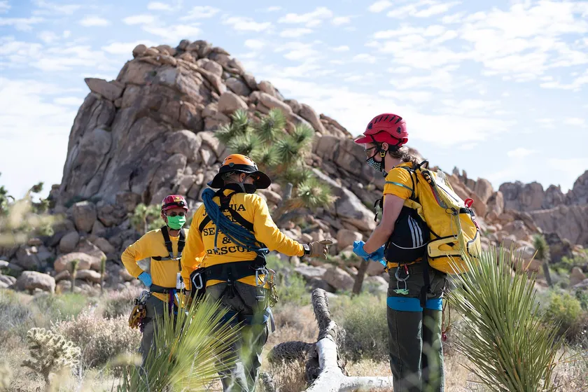 Three search-and-rescue team members in yellow shirts and helmets stand with coils of rope in rocky desert