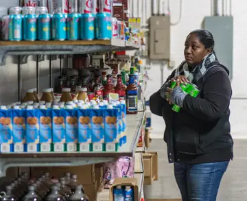 Two women in a food-bank warehouse packing canned goods into cardboard boxes