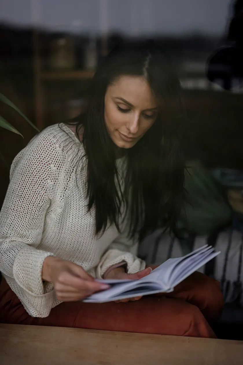 A woman in a cream sweater sitting by a window and reading an open book
