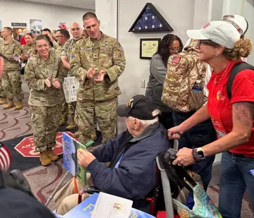 Service members in camouflage clap as a woman pushes an older veteran in a wheelchair past a small crowd