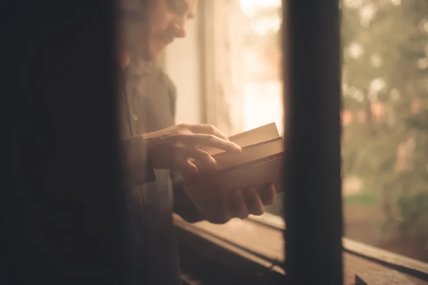 An adult by a bright window, holding an open book and turning a page