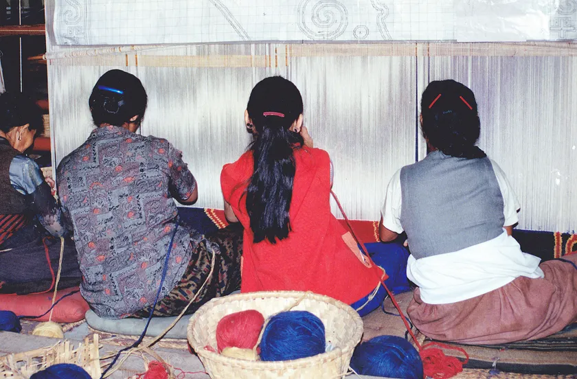 Three women working at a large wooden loom, pulling threads through the weave