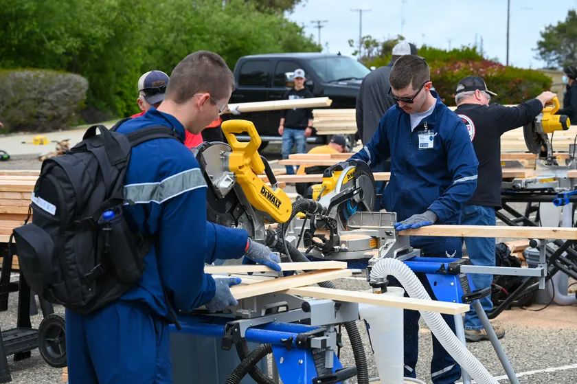Adults cutting lumber at outdoor tables during a bed-building event