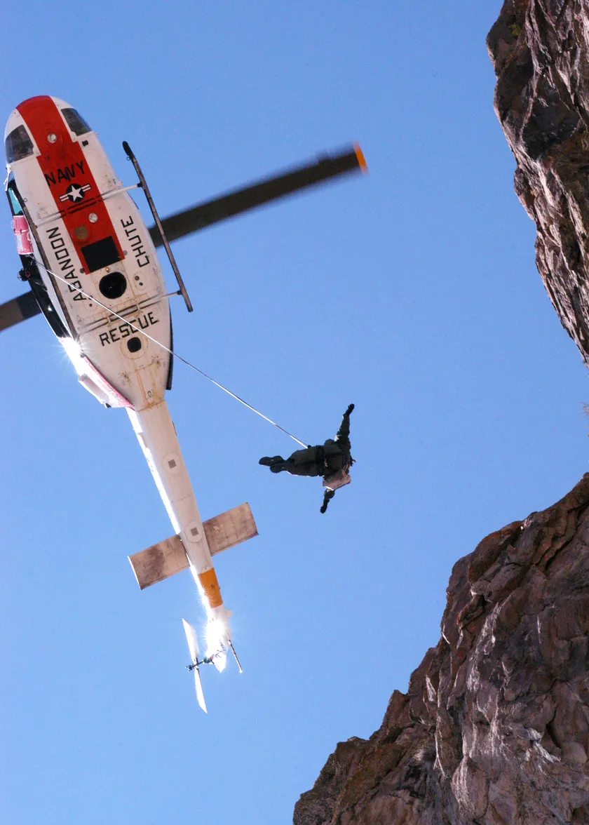 A rescuer hangs from a cable under a white and red Navy rescue helicopter beside a rock cliff