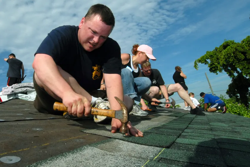 A worker in a blue uniform on a roof, nailing shingles into place