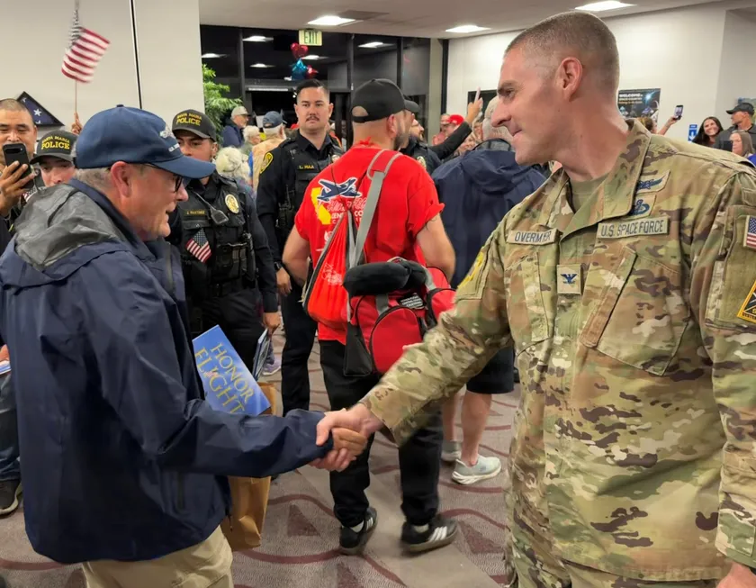 A service member in camouflage shakes hands with an older man holding a blue Honor Flight folder