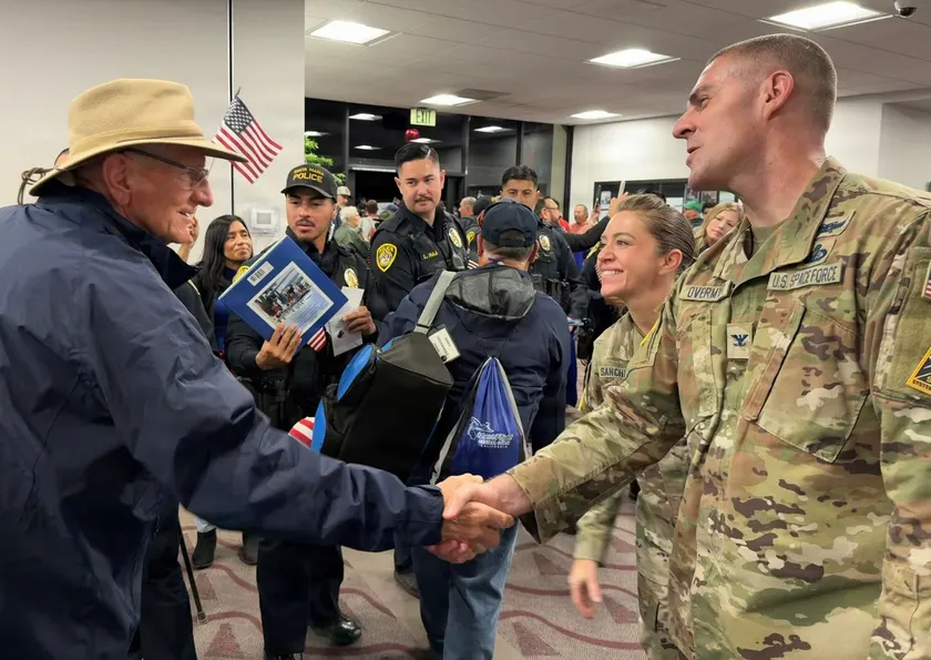 A service member in camouflage shaking hands with a veteran in a hat at an airport welcome