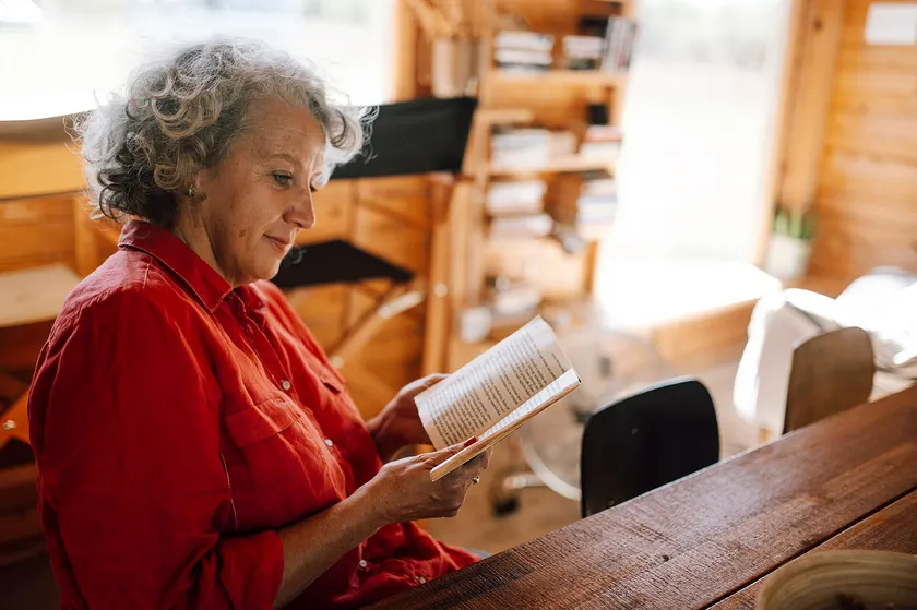 An older woman in a red shirt reading an open book at a wooden table