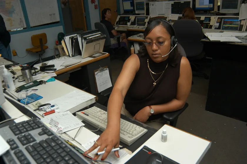 A woman wearing a telephone headset works at a desk of phones and computer screens in a 911 center