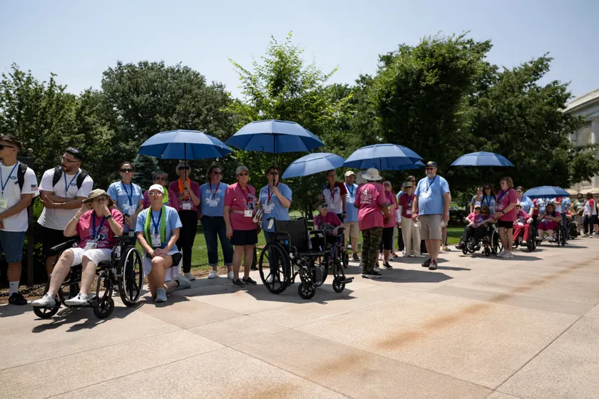 Veterans and companions with blue umbrellas gathered on a stone plaza beside a white memorial building